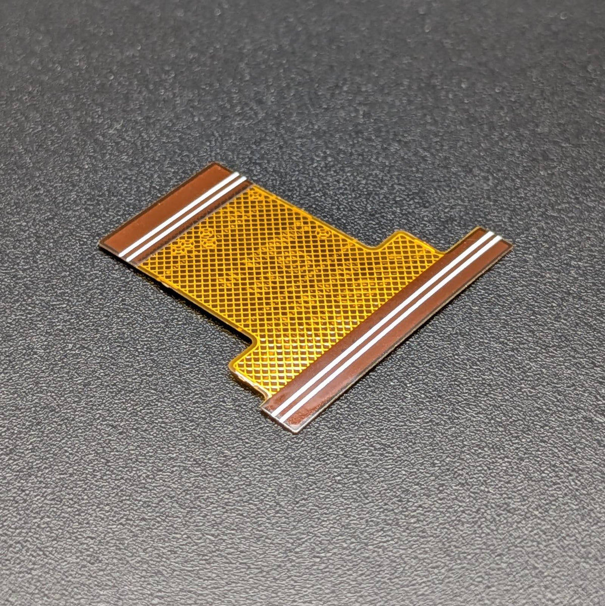 A small flexible circuitry connector piece with a gold mesh center and brown copper traces, photographed on a dark textured surface.