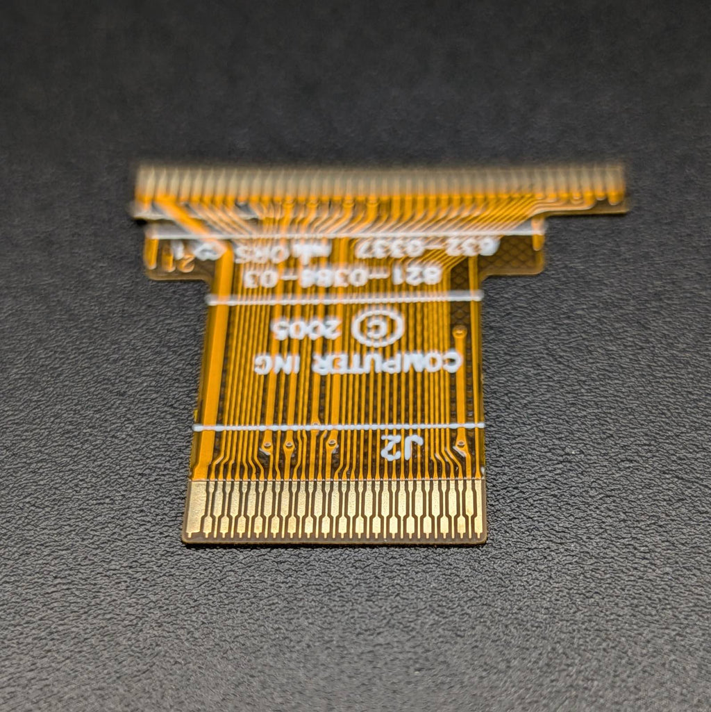 A small gold flexible circuit board connector with white printed text and markings, photographed on a dark textured surface.