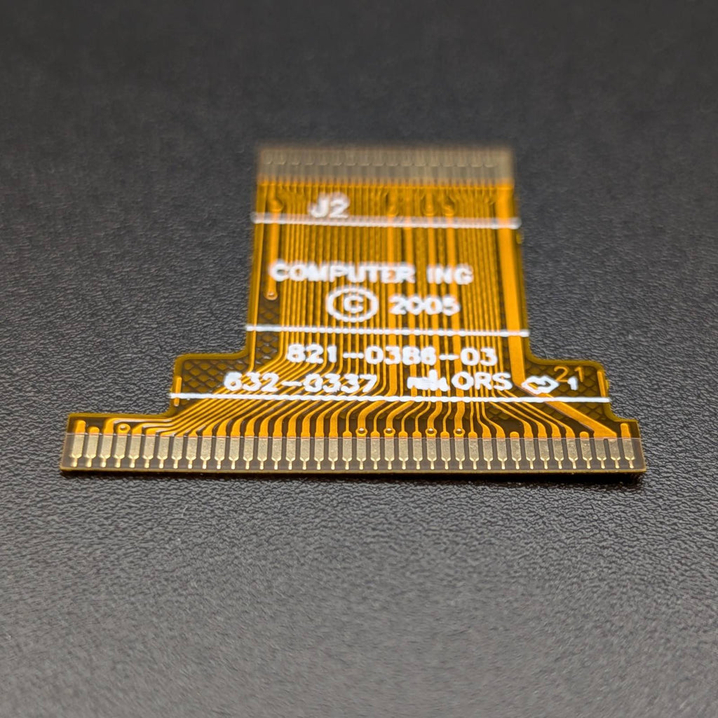 A small gold flexible circuitry connector with white printed text reading 'COMPUTER INC 2005' and pin contacts along the bottom, photographed on a dark textured surface.