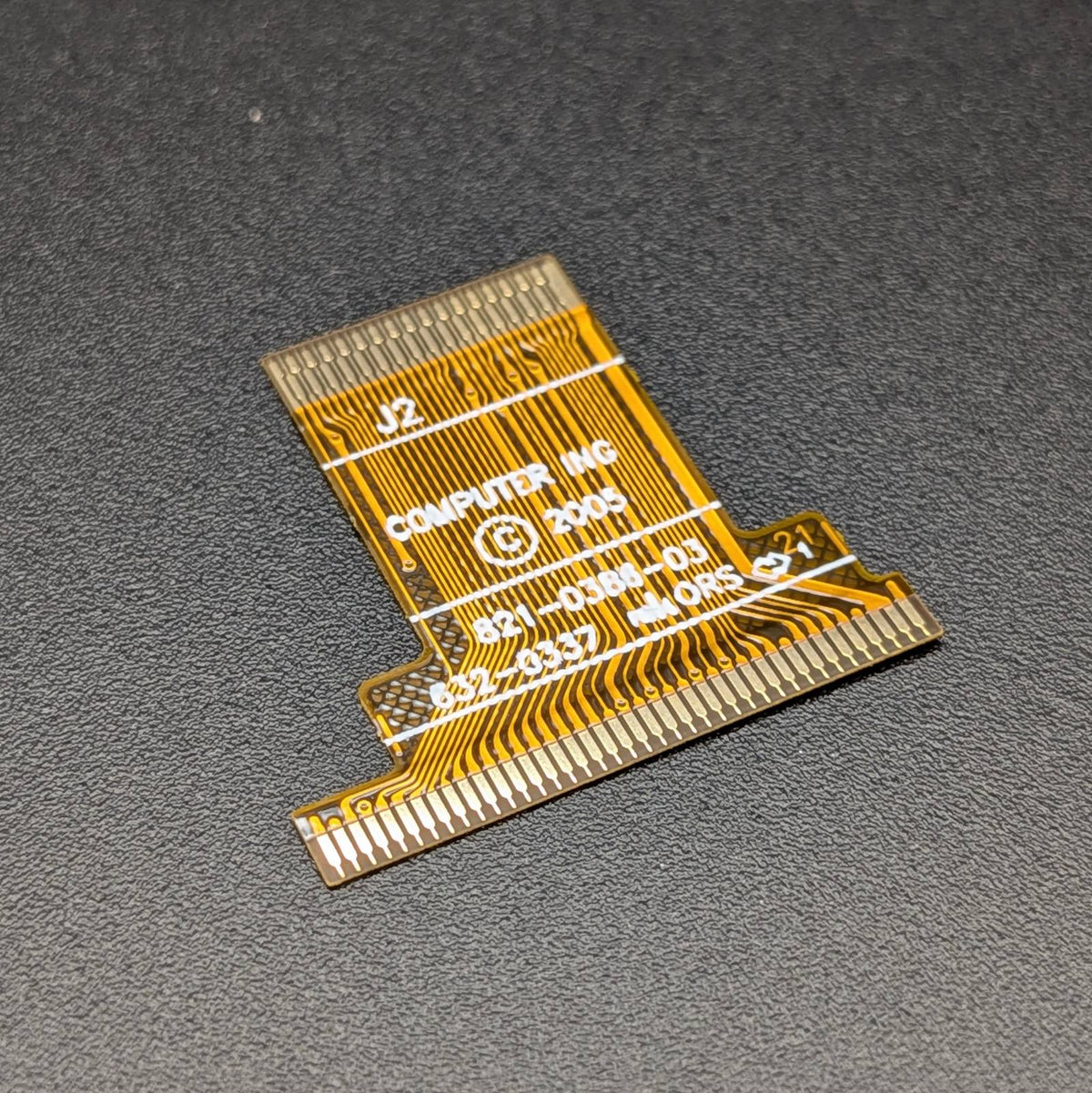 A small gold flexible circuitry connector board with white printed text and markings, photographed on a dark textured gray background.