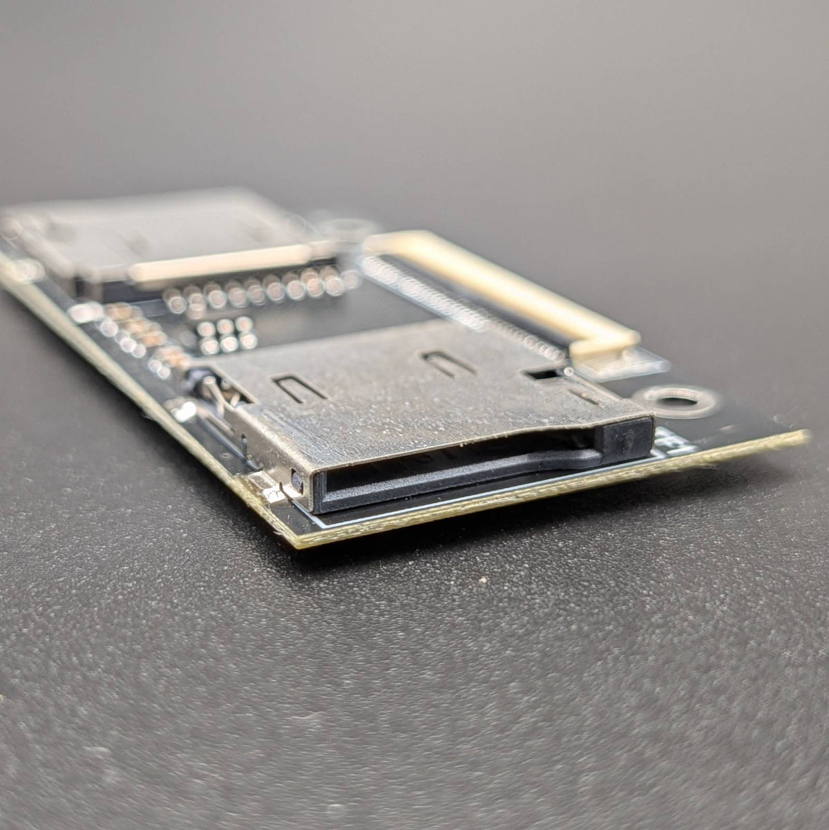 A close-up of a small electronic circuit board module with a black plastic connector and gold-toned metal contacts, photographed on a dark gray textured surface.