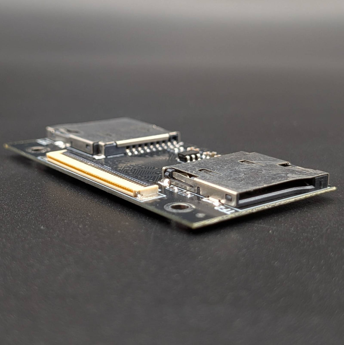 A small electronic circuit board module with two metal connector slots, photographed on a dark gray surface.