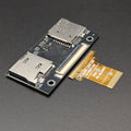 A small electronic adapter board with two silver metal connector ports on a dark circuit board, shown next to a gold flexible ribbon cable on a dark textured background.