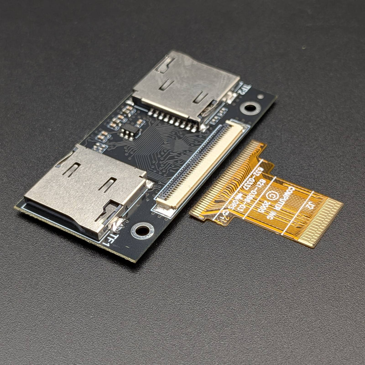 A small electronic adapter board with two silver metal connector ports on a dark circuit board, shown next to a gold flexible ribbon cable on a dark textured background.