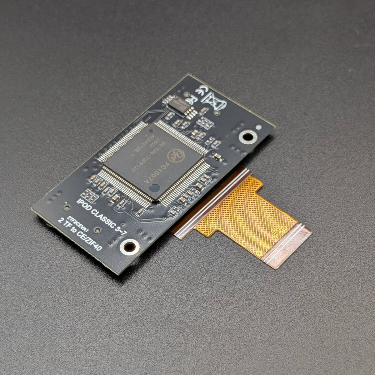 A small electronic circuit board with a large integrated circuit chip and a gold flexible flat cable, photographed on a dark textured surface.