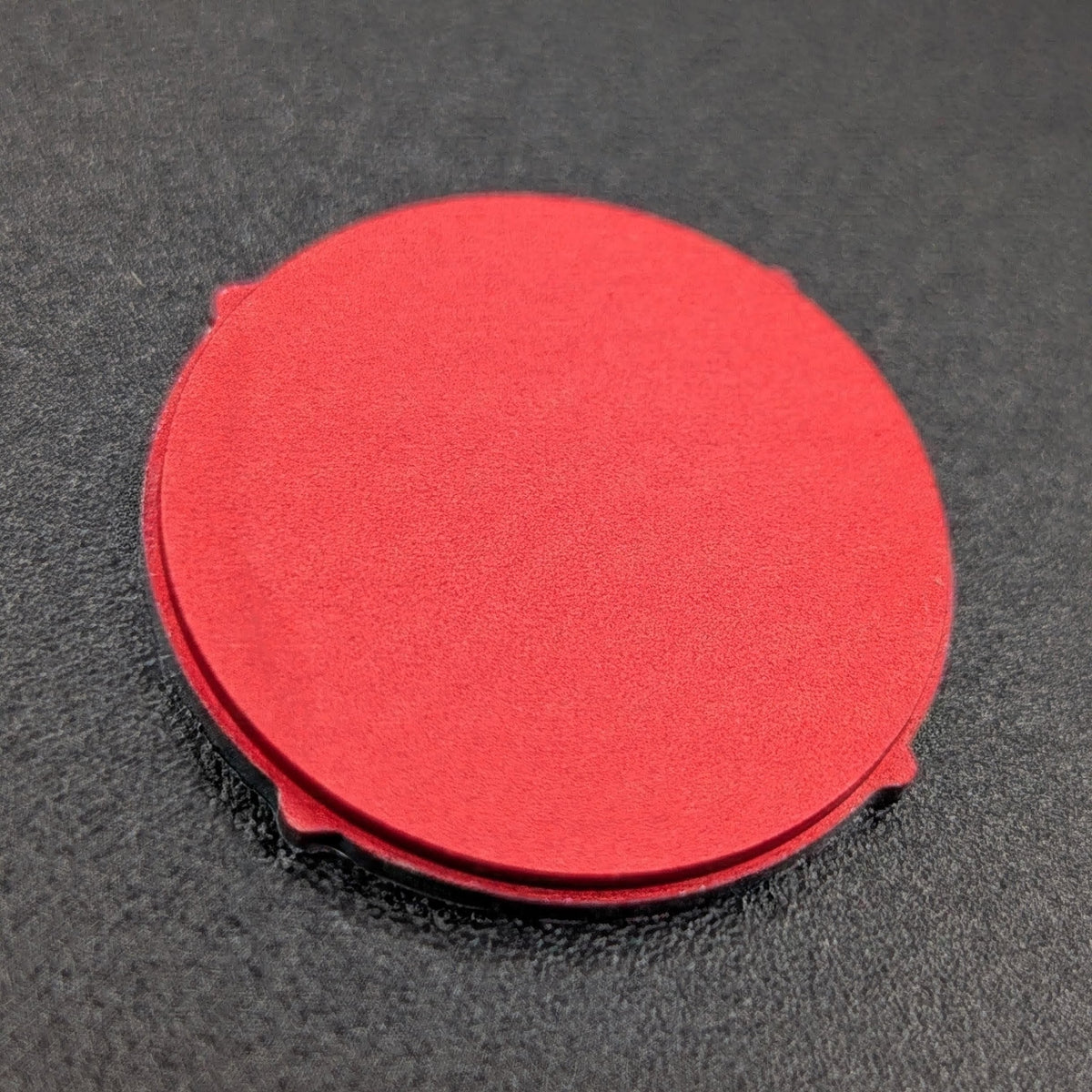 A red button with small tabs around the edges, photographed on a dark textured surface.