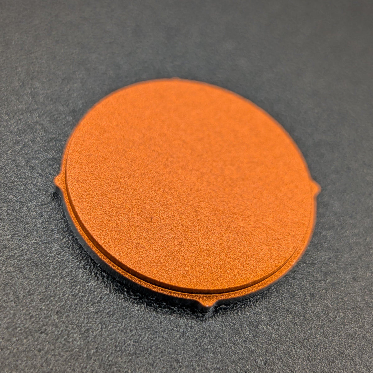 A round orange textured button with small tabs on the sides, photographed on a dark gray textured surface.
