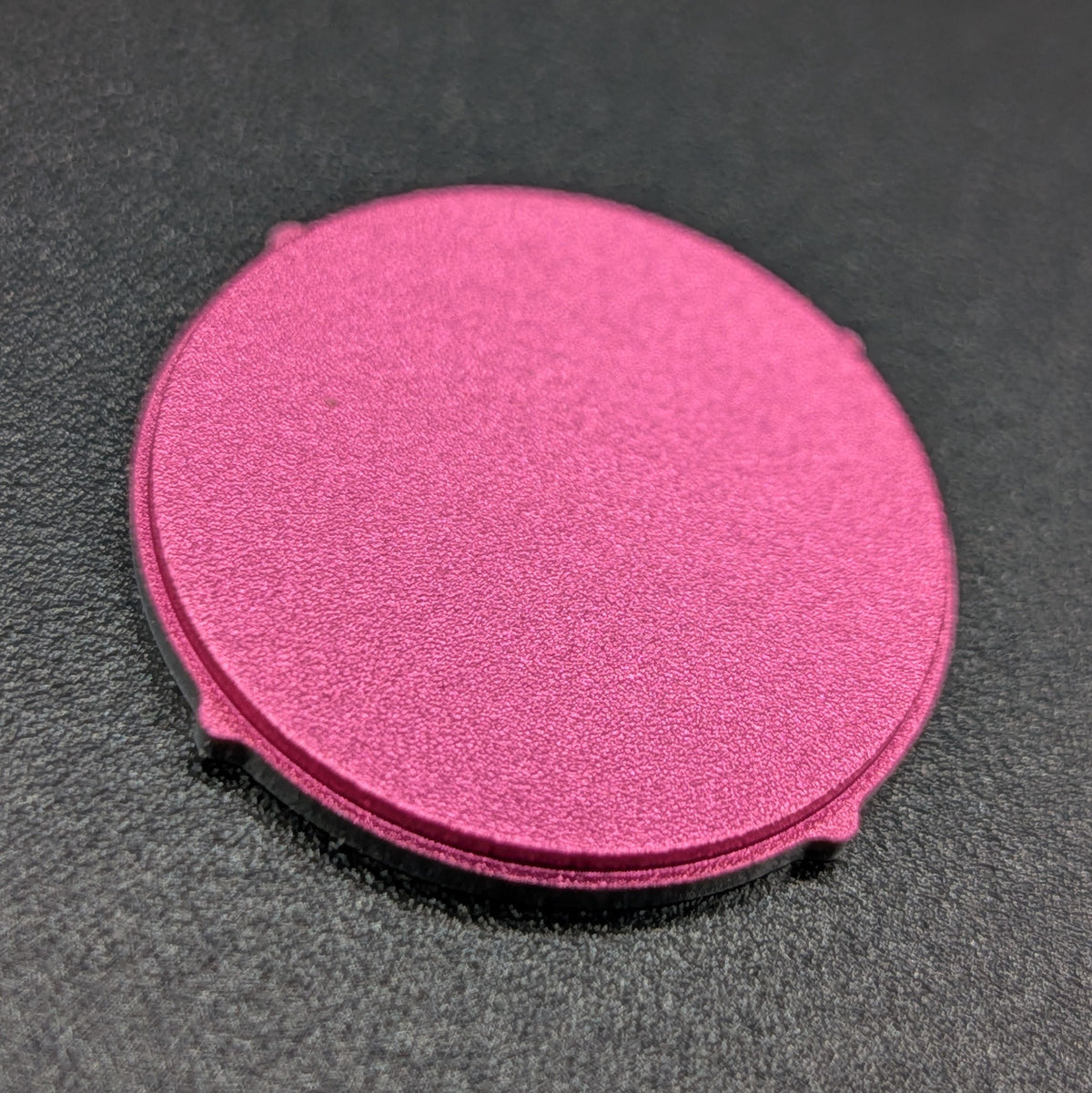 A circular, textured metallic pink component with small tabs on its outer edge, resting on a dark gray textured surface.