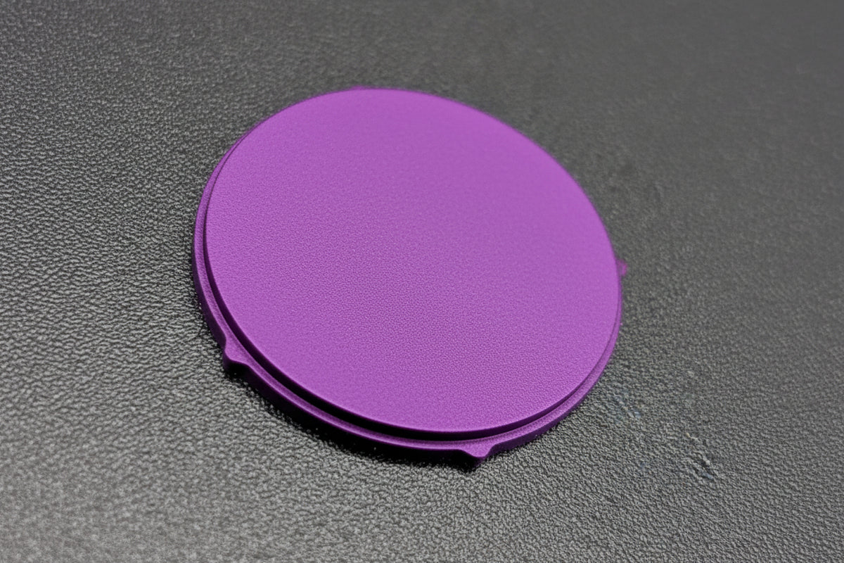 A round, flat button with small tabs on the sides, photographed on a dark textured surface.
