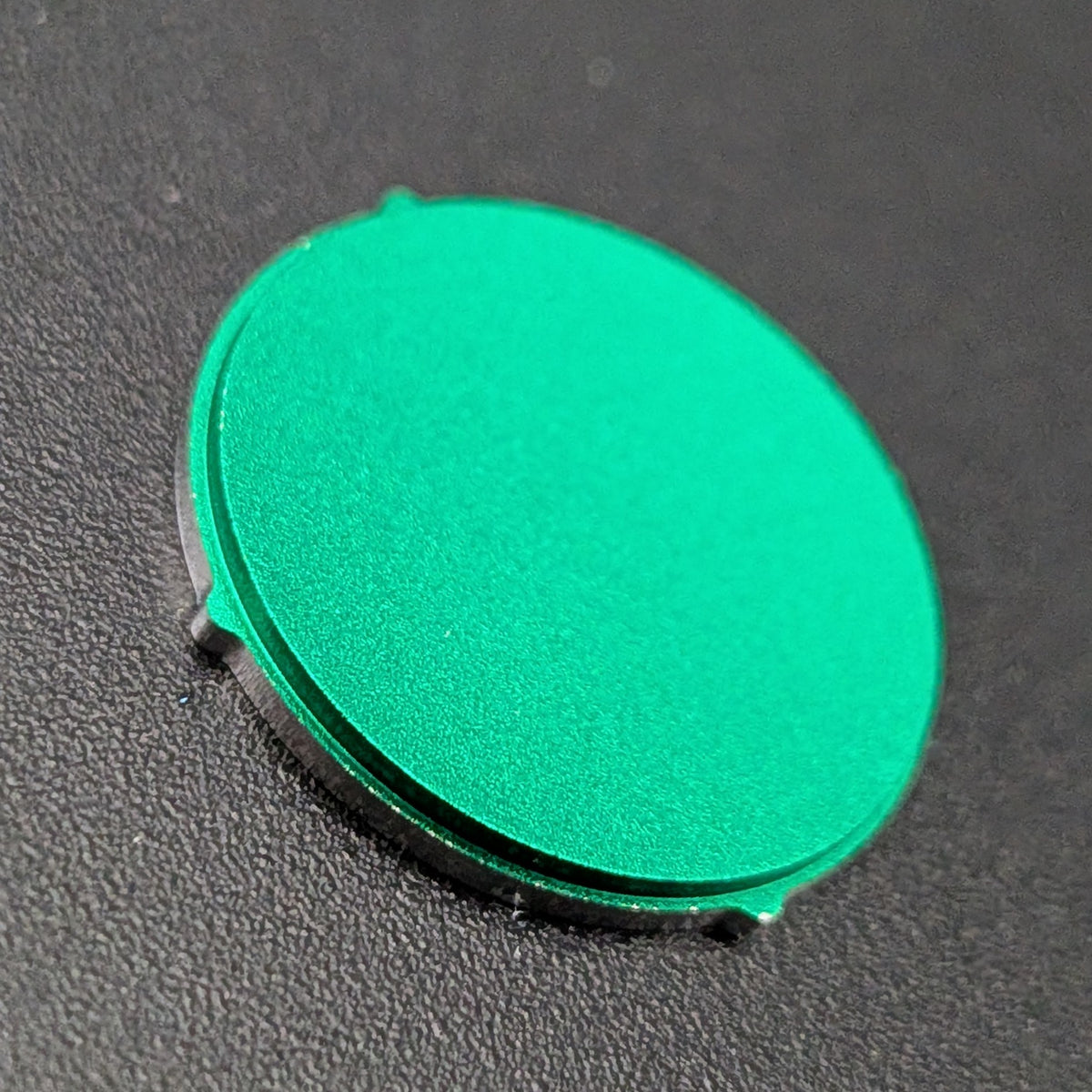 A round green metallic button with a matte finish, resting on a dark textured surface.