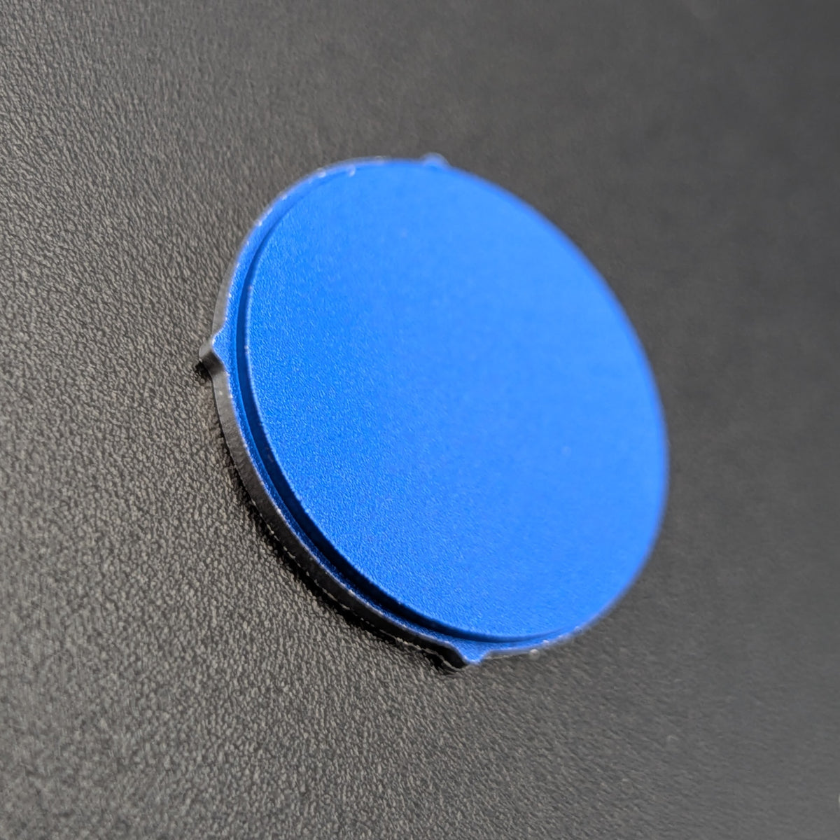 A round blue button, shown on a textured dark gray surface.