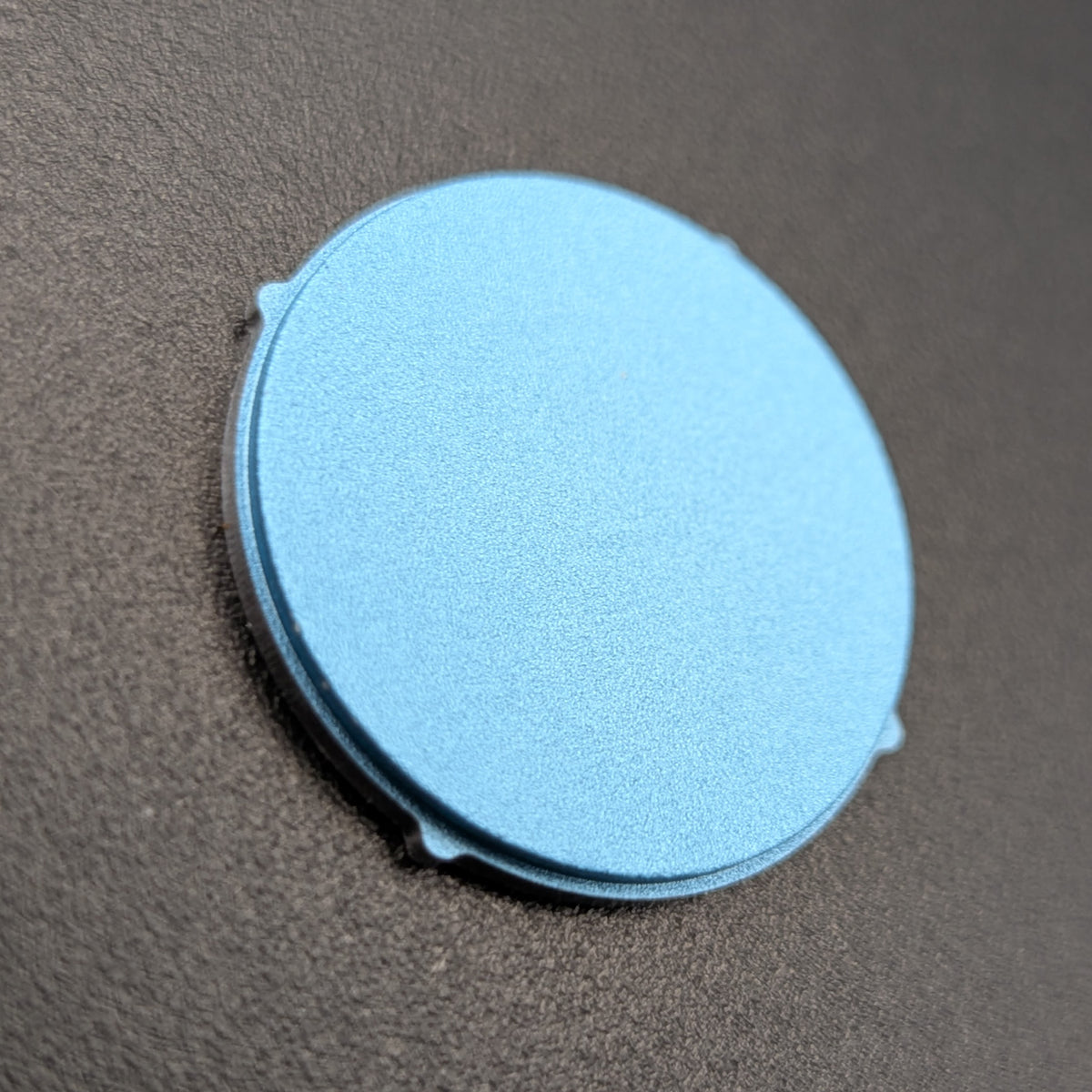 A round, light blue textured disc with small tabs on the sides, resting on a dark grey textured surface.