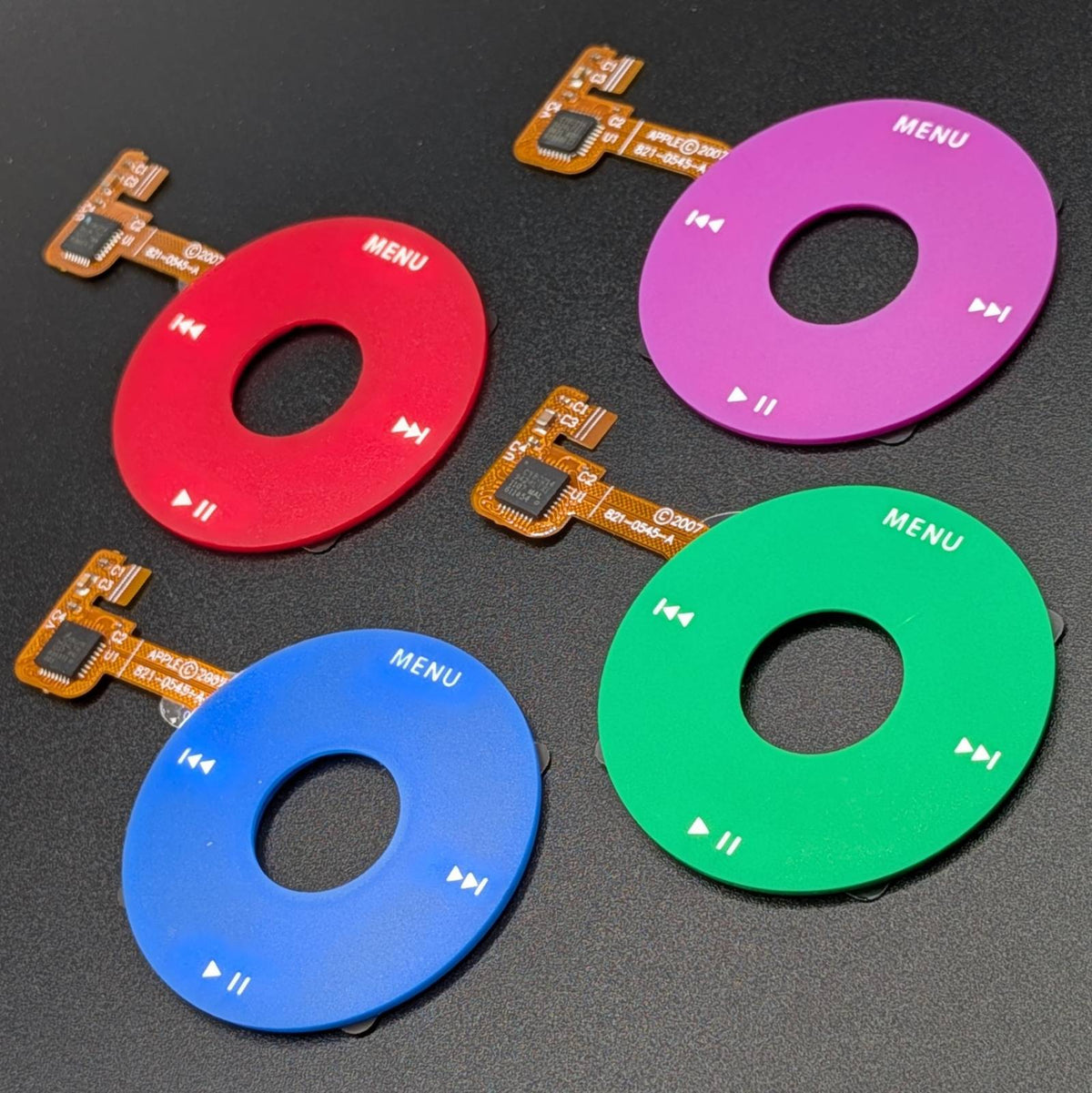 Four round electronic control buttons in red, purple, blue, and green, each with a center cutout and attached flex cable, arranged on a dark textured background.