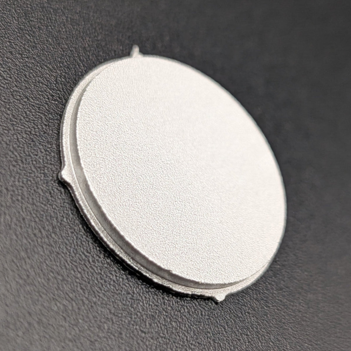 A round, flat silver metal disc with small tabs on the side, photographed on a dark gray textured background.